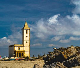 La silueta de la iglesia de San Miguel se alza como un símbolo de resistencia frente al mar y las salinas del Cabo de Gata. Vista de la iglesia de San Miguel en la Almadraba de Monteleva, Cabo de Gata. Un edificio religioso blanco y solitario con un campanario sencillo, situado en un paisaje llano y costero bajo un cielo despejado.
