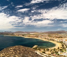 La unión perfecta entre el urbanismo costero y la serenidad del mar bajo un cielo infinito Paisaje panorámico de una costa urbana con playas de arena dorada y mar azul. En el fondo se ven montañas bajas y marrones bajo un cielo azul lleno de nubes blancas y delgadas. En primer plano, la ladera de una colina con vegetación escasa domina el ángulo inferior izquierdo.