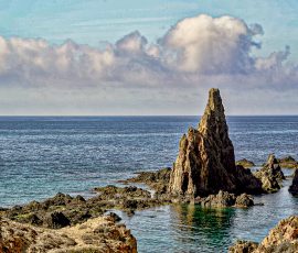Una formación rocosa vertical desafía un cielo de nubes algodonosas. Una gran formación rocosa vertical y puntiaguda en medio del mar azul. El agua es transparente cerca de la base de la roca, revelando tonos verdosos. Al fondo, una banda de nubes blancas horizontales cubre parte del horizonte bajo un cielo azul.