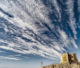 Una antigua torre de vigilancia se mantiene firme frente a un despliegue de nubes cirros que parece cobrar vida propia. Una torre de piedra histórica situada en una colina bajo un cielo azul vibrante lleno de nubes blancas largas y estriadas que cruzan diagonalmente la imagen. La torre tiene ventanas pequeñas y parece antigua. Una valla de madera recorre el borde de la colina.