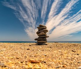 Una columna de piedras en equilibrio perfecto desafía la inmensidad del horizonte. Primer plano de varias piedras planas apiladas verticalmente en una playa desierta. Al fondo, el mar azul bajo un cielo brillante donde nubes blancas alargadas irradian desde el centro hacia arriba, enmarcando la escultura de piedra.