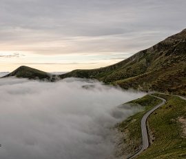 Una serpenteante carretera de montaña se asoma a un océano de nubes al atardecer. Una carretera con curvas cerradas recorre la ladera de una montaña verde y escarpada. Debajo de la carretera, un mar de nubes blancas muy densas cubre todo el paisaje inferior. Al fondo, se ven picos de montañas lejanas bajo un cielo gris claro con luz de atardecer.