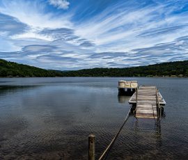 La estructura de madera guía la mirada hacia la serenidad de la bahía. Perspectiva desde el inicio de un largo embarcadero de madera que se adentra en un mar calmado. Las tablas de madera del suelo crean líneas que dirigen la vista hacia el horizonte. El agua es de un azul claro y el cielo está prácticamente despejado.