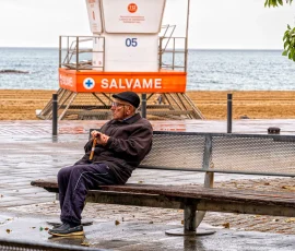 A veces el mayor grito es el que nadie escucha. Anciano con bastón en un banco del paseo marítimo, con suelo mojado y una caseta de salvamento detrás.