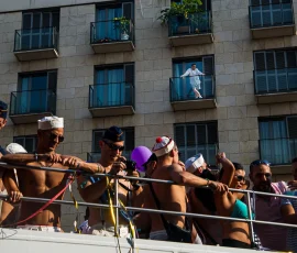 Mientras un grupo de personas se lo están pasando en grande encima de un autocar descubierto a pleno Sol, un hombre, en un edificio observa, sin aparente interés, lo que sucede en la calle. Hombre en un balcón observando un desfile de personas a pecho descubierto encima de un autocar.